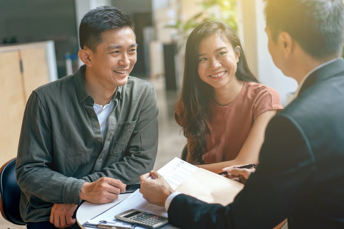 Two people smile while sitting at a table with another person wearing a suit and holding documents. 