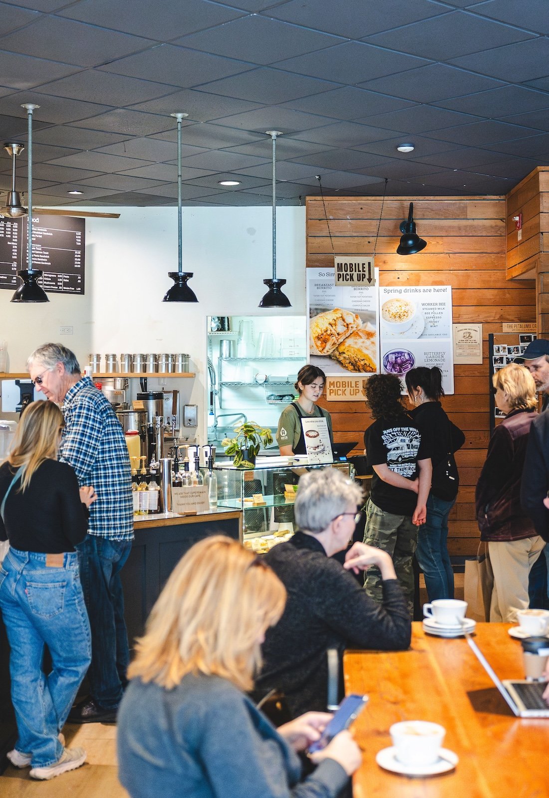 Customers in a busy coffee shop. In the foreground, some sit at a wooden table. In the background, a customer orders from a barista. The room is brightly lit.