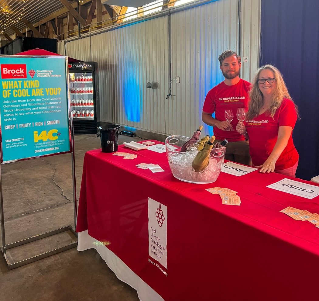 A man and a woman stand behind a table that holds various flyers and a tub holding wine bottles. Both are wearing red shirts.