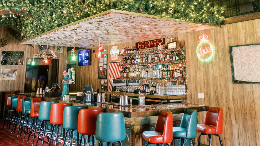 A festive bar interior decorated with lush green garlands and colorful Christmas lights. The bar has a warm, cozy atmosphere with red and green upholstered bar stools lined up along a polished wooden counter. Above the bar, a large digital countdown clock is displayed, and a neon sign reading 'Lala’s' is illuminated on the wood-paneled wall. The ceiling features exposed wooden beams adorned with string lights and a rustic chandelier, contributing to the holiday ambiance.