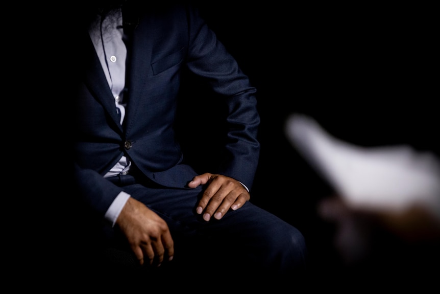 A person wearing a suit is sitting in a chair in a dark room, their face is not visible.