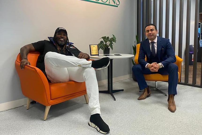 Cricketer Chris Gayle sits in an office chair and smiles next to Melbourne businessman Amit Miglani.