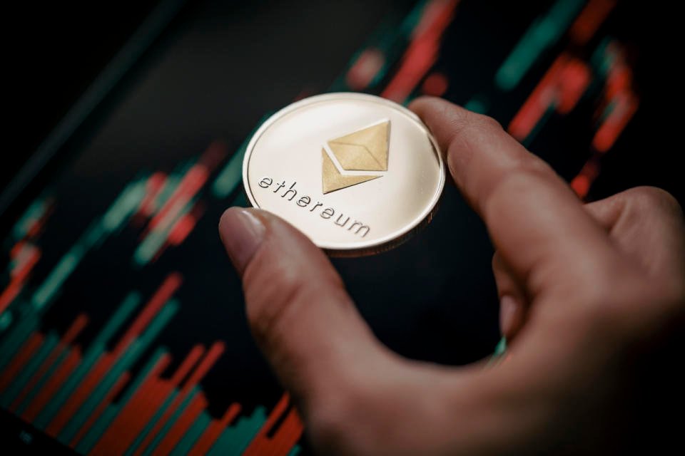 Hand holding a silver Ethereum (ETH) cryptocurrency coin with candle stick graph chart and digital background.