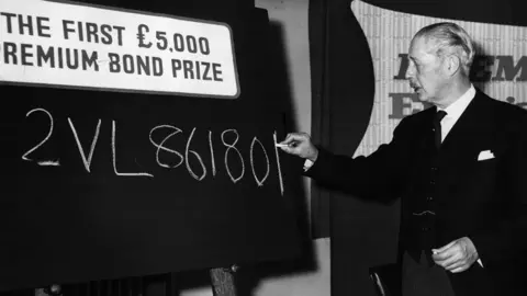 Fox Photos/Getty Images A black and white image of Prime Minister Harold MacMillan (1894-1986) writing the number of the first ever £5,000 Premium Bond prize on a chalkboard  at the National Savings headquarters in London.