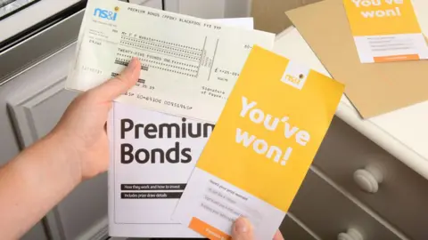 Alamy A person's hands holding Premium Bonds literature, including a leaflet saying they have won a prize and a cheque for £25. Part of a white door with a silver letterbox is to the left and a light brown envelope and another NS&I leaflet are on top of a set of drawers to the right