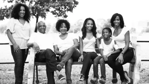 Melanie Clarke A black and white photograph showing smiling family members, from left to right: Melanie Clarke, Hugo Clarke (her father), Margaret Clarke (Melanie's mother), Danielle (Melanie's niece), Daelen (Melanie's niece) and Gillian (Melanie's sister). Melanie is standing beside a bench that everyone else is sitting on. They are all wearing light coloured tops and dark trousers or jeans. Picture taken in 2013