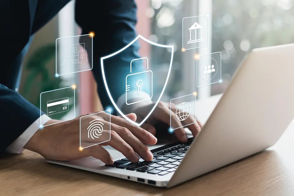 A person typing on a laptop with digital security icons floating above the keyboard, including symbols for a shield, lock, fingerprint and online banking, representing data protection and cybersecurity.
