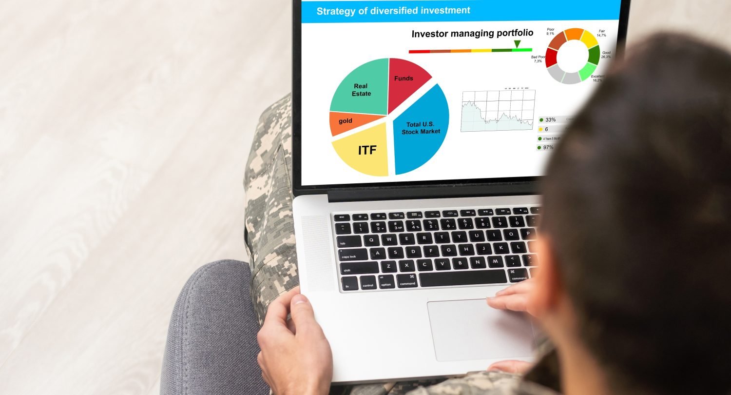 A person wearing camouflage pants and a grey shirt sits holding a silver laptop on their lap, viewed from a slight overhead angle from behind their right shoulder. The laptop screen displays a financial dashboard titled 'Strategy of diversified investment.' The screen features a large pie chart showing asset allocations for 'Total U.S. Stock Market,' 'Funds,' 'Real Estate,' 'gold,' and 'ITF.' A smaller donut chart indicates portfolio performance categories with percentages, and a line graph shows market trends. The person's right hand rests on the laptop's trackpad. A light wooden floor is visible in the background.