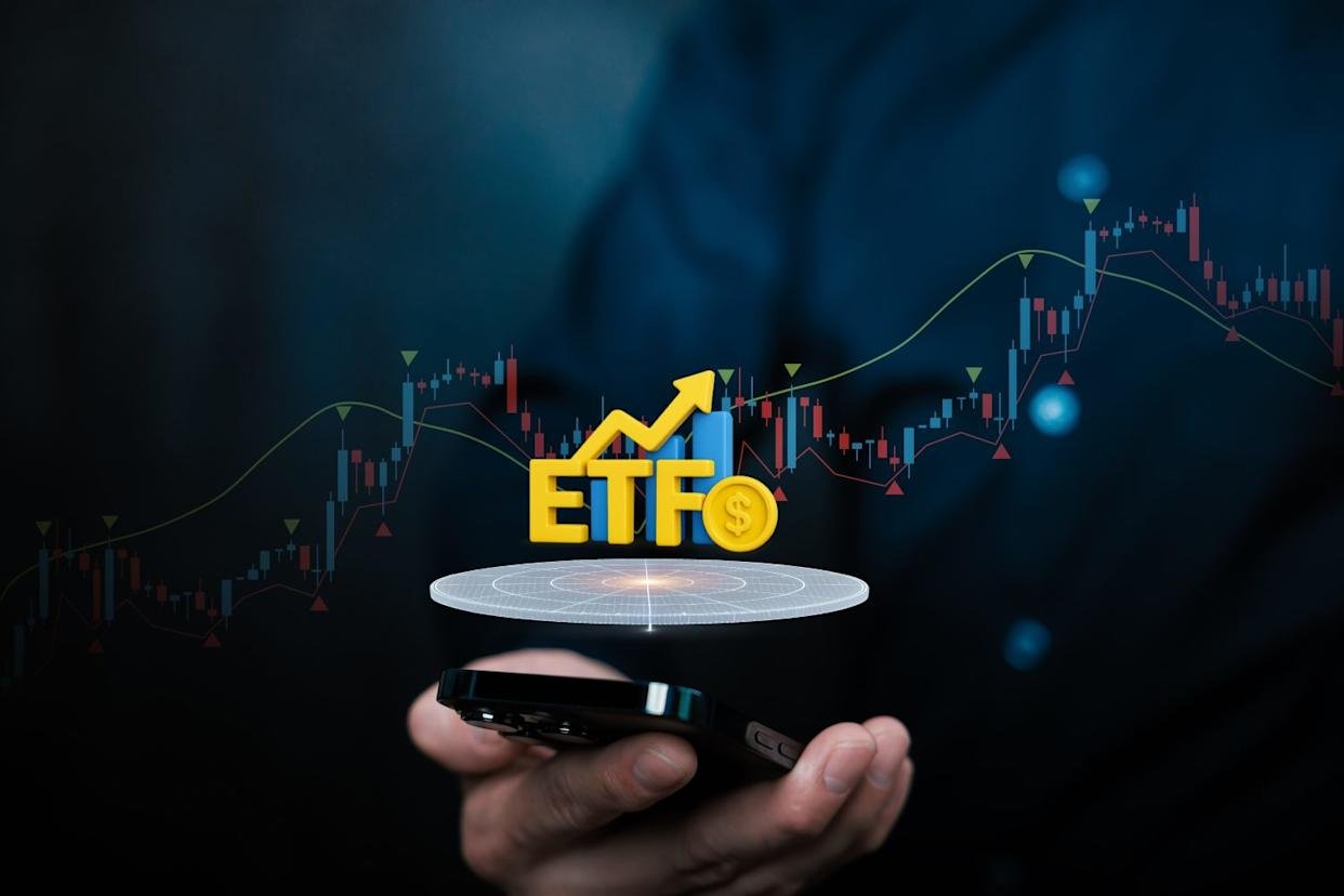A close-up of a person holding a black smartphone, with a glowing circular holographic platform hovering above it. On the platform, yellow 3D letters spell 'ETFO', accompanied by a dollar sign coin, an upward-pointing arrow, and blue bar graphs, all signifying financial growth. A blurred stock market candlestick chart with red and blue candles forms the background.