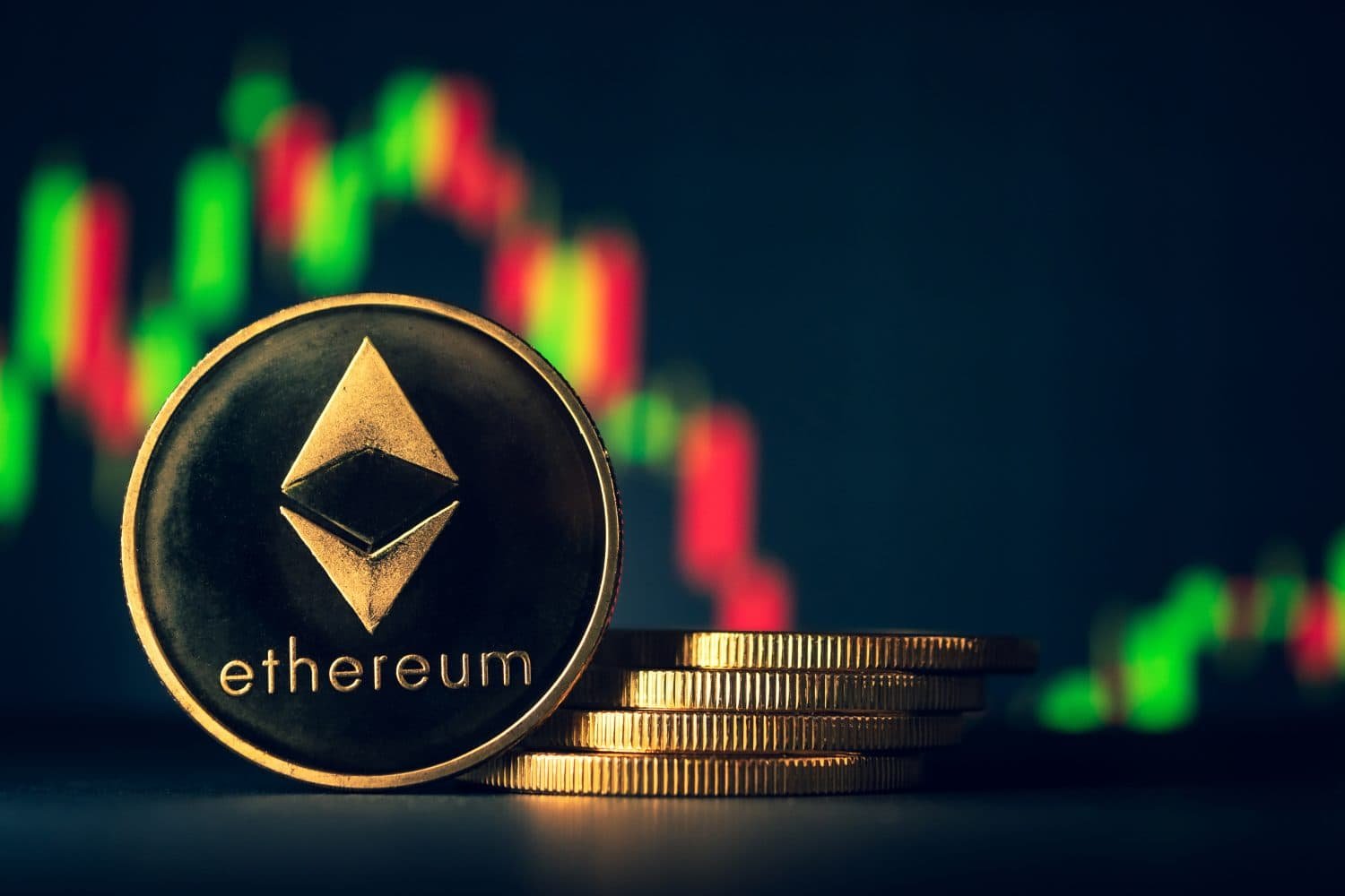 A close-up shot of a physical gold and black Ethereum coin in the foreground, with the word 'ethereum' and its geometric symbol visible. In the blurred background, red and green market charts indicate financial trends, and a stack of golden coins is partially visible to the right of the main Ethereum coin.