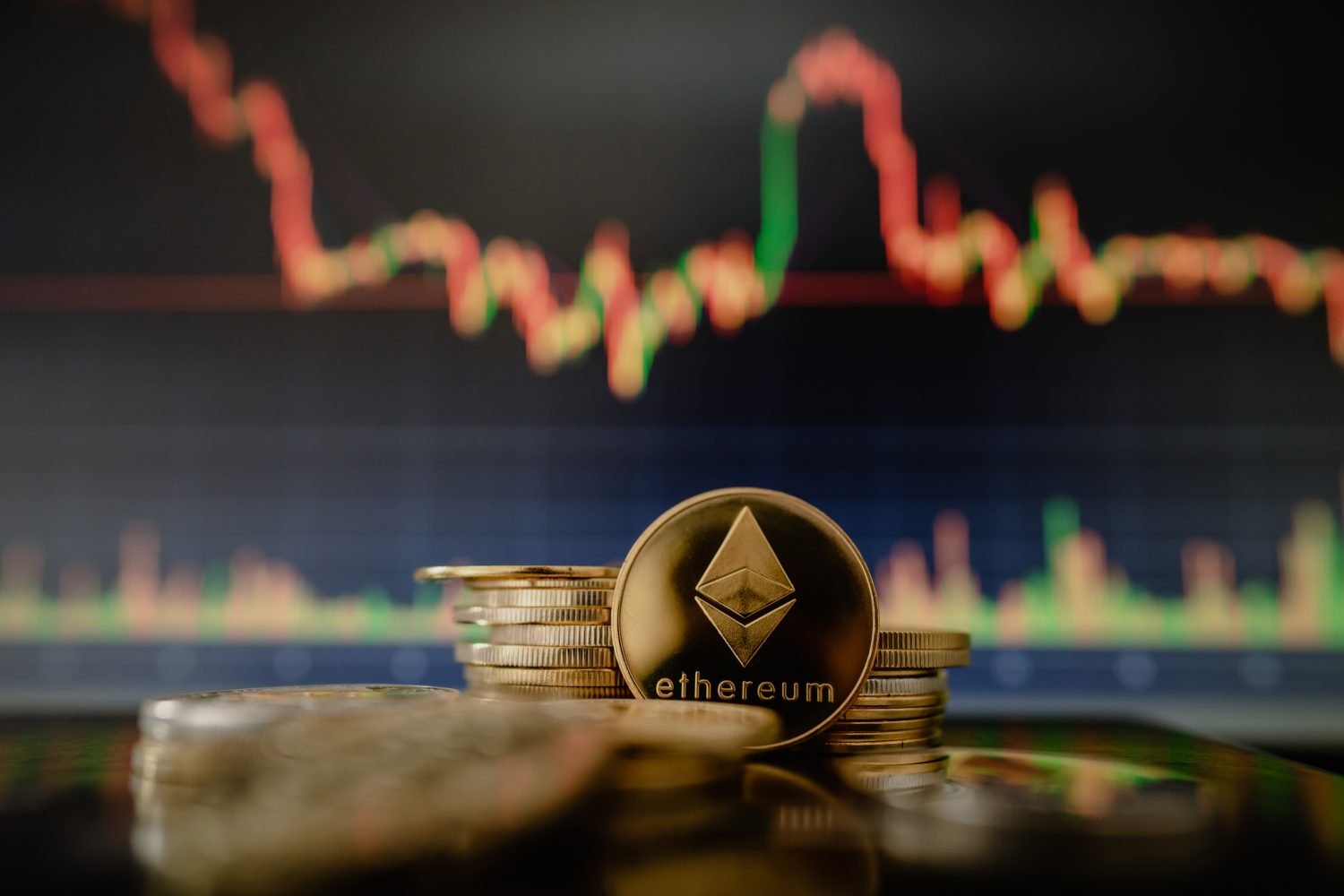 A close-up of a golden Ethereum coin, featuring the Ethereum logo and 'ethereum' text, stacked among other gold-colored coins. In the out-of-focus background, a digital screen displays a financial market chart with fluctuating red and green lines, indicating price changes.