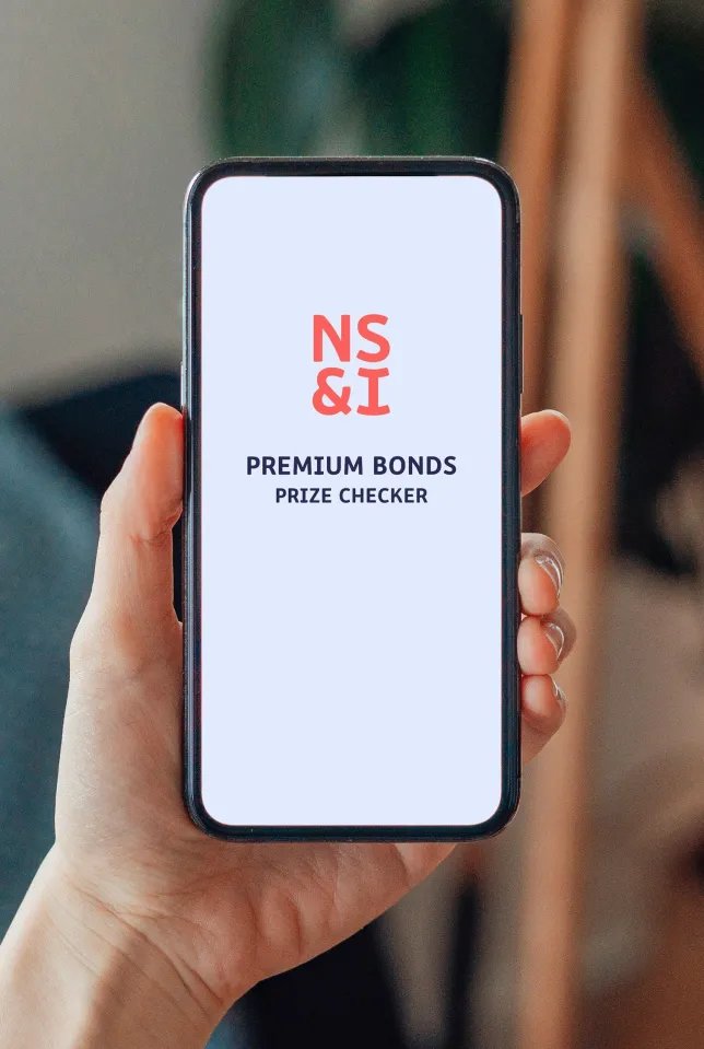 A hand holds a smartphone displaying the NS&I Premium Bonds Prize Checker app.