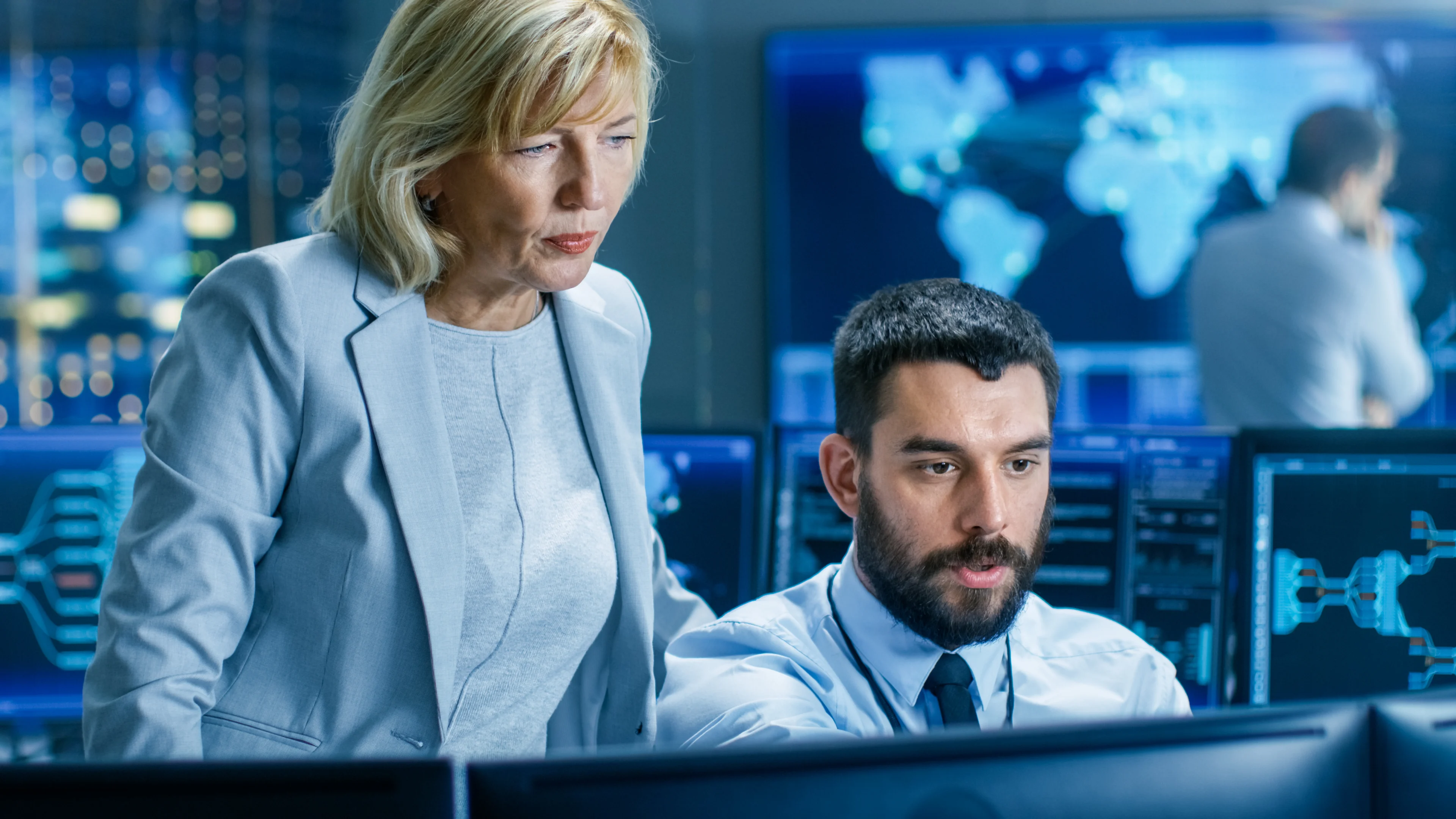 Two traders, one standing, one sitting, look at data on screens in an office. 