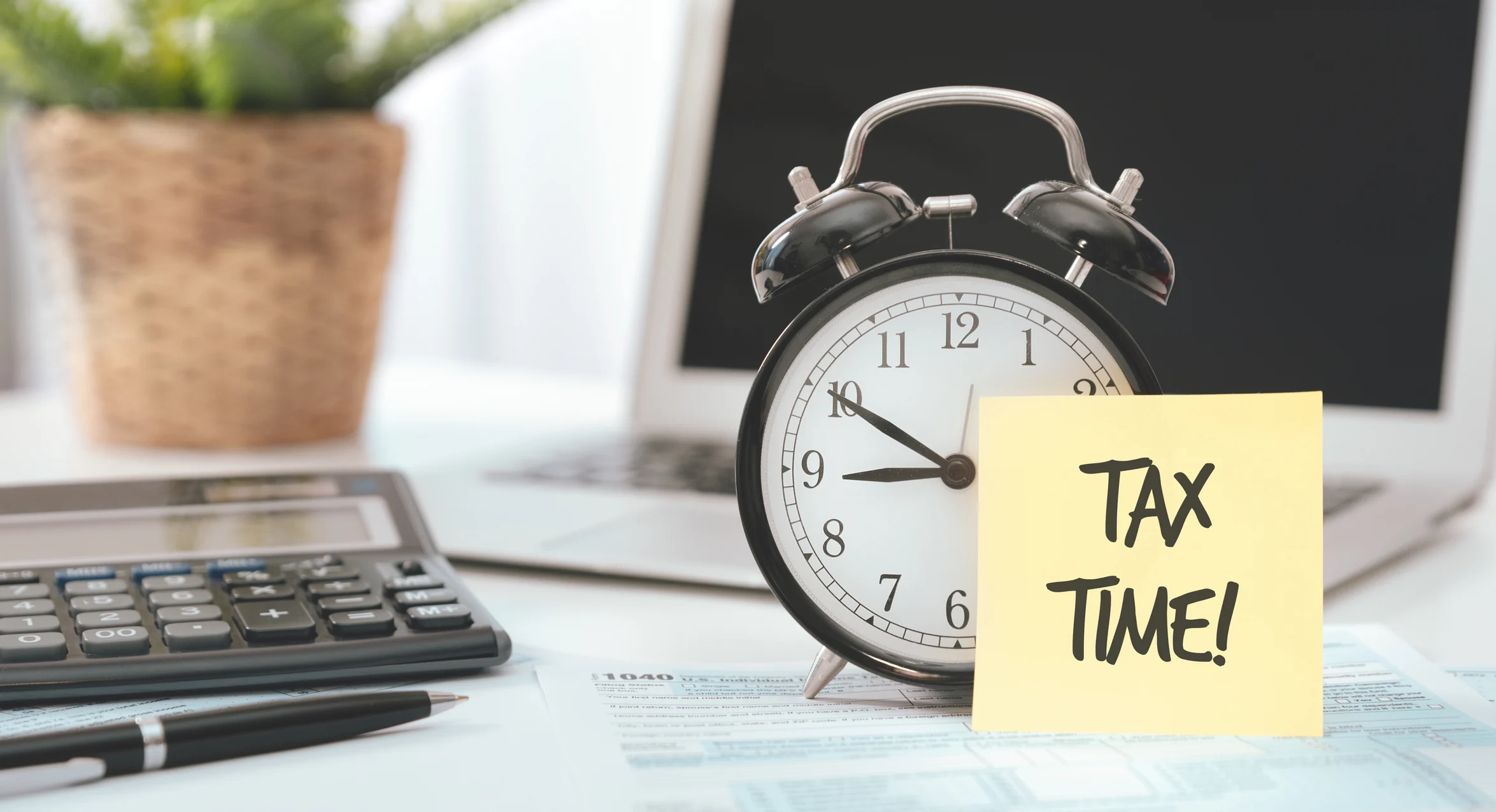 A desk, with a plant, open laptop, calculator, pen, and alarm clock on it. Propped against the clock is a yellow note reading, "Tax Time!"