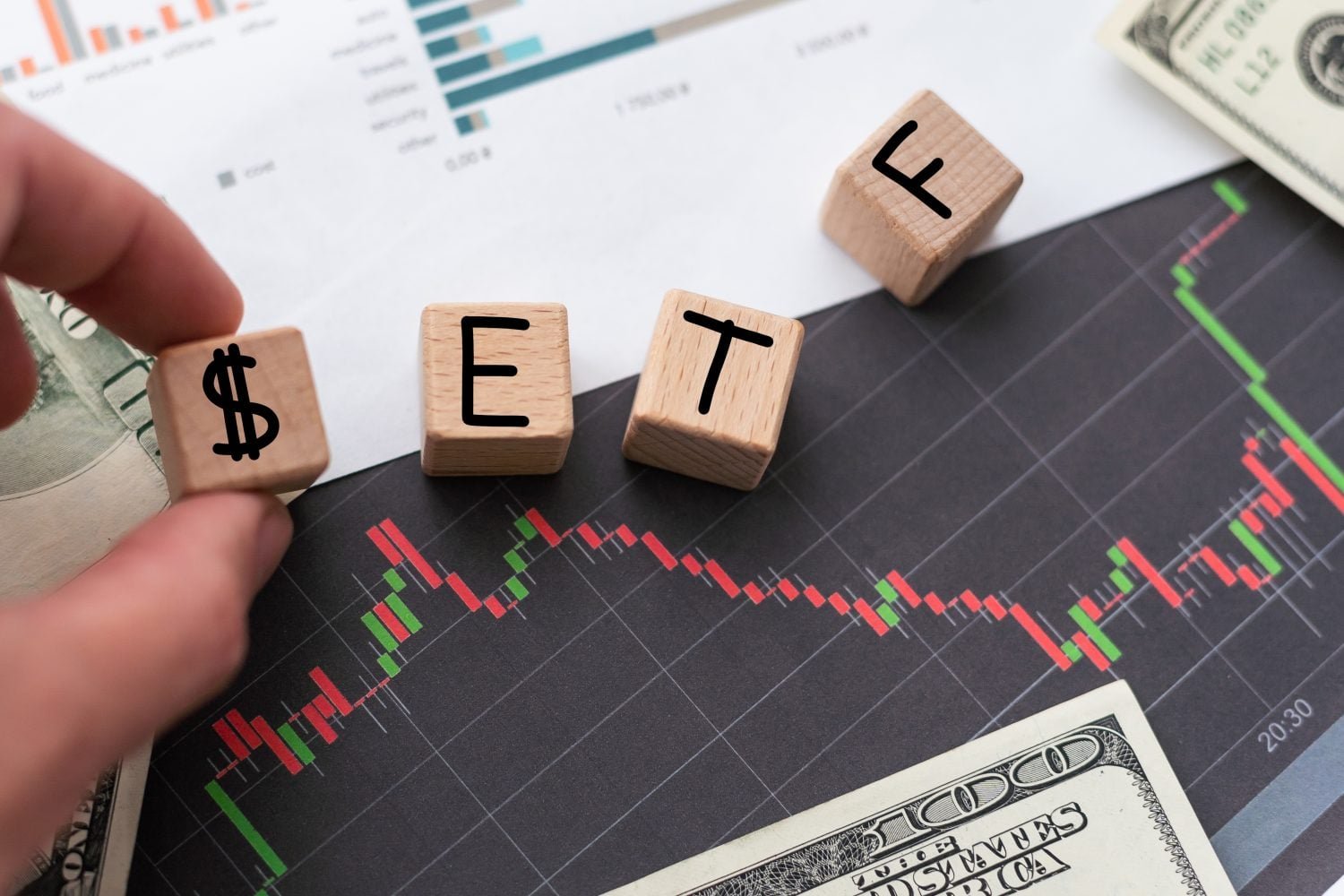 A close-up shot of a hand placing a wooden block with a dollar sign ($) on a black financial chart. Other wooden blocks spell out 'ETF'. The chart shows a line graph with green and red candlestick patterns indicating market movements. US dollar bills are visible in the top right and bottom right corners of the image, and a white paper with bar graphs is in the background.
