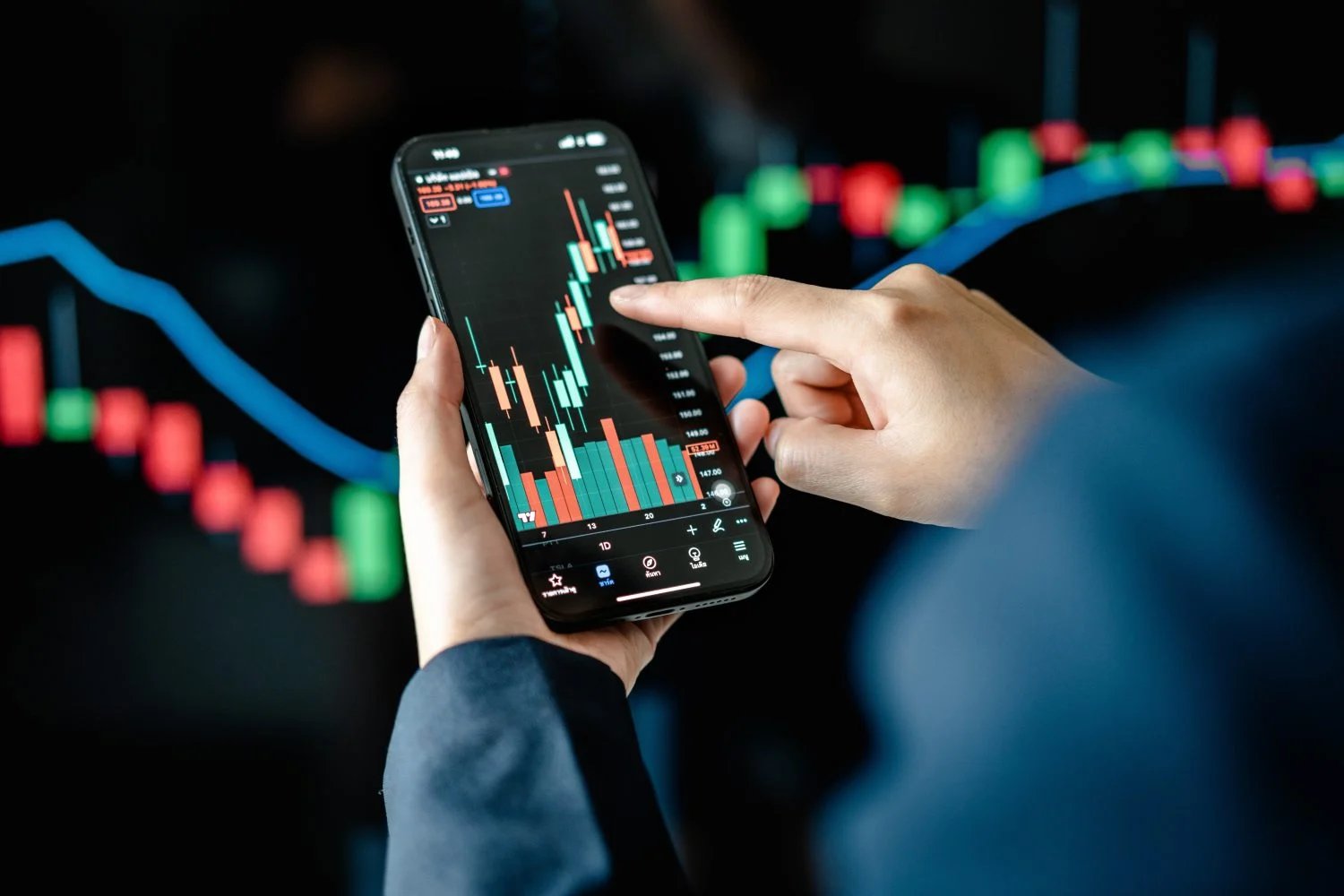 A close-up shot shows a person holding a black smartphone horizontally, displaying a vibrant green and red candlestick stock chart against a dark background. A finger from the right hand is pointing to a specific point on the chart. In the blurred background, another larger screen displays a similar financial chart with a prominent blue trend line and red and green vertical bars.