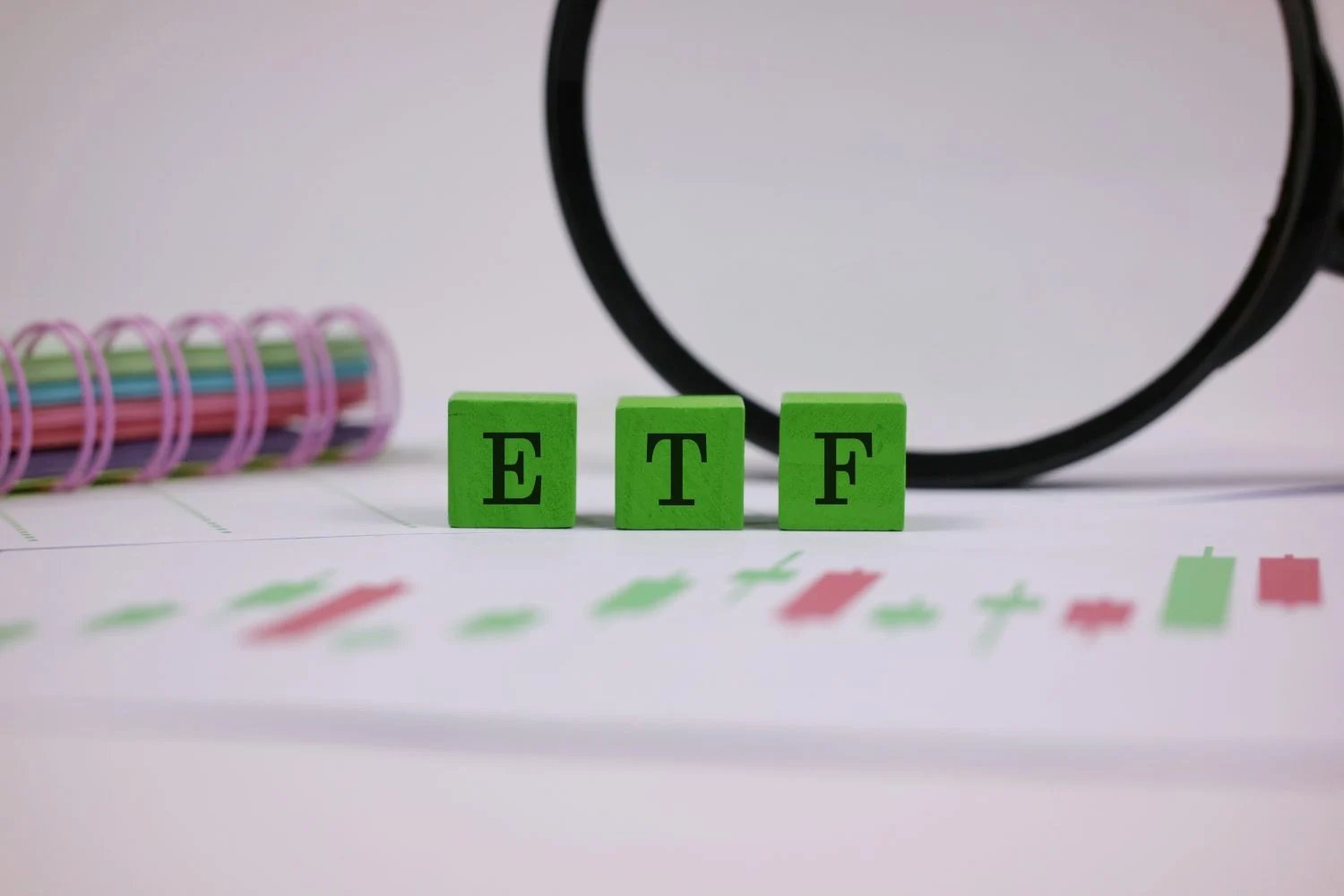 Three green wooden blocks with black letters spell 'ETF' in the foreground. They rest on a white document showing a blurred financial chart with red and green candlesticks. A large magnifying glass with a black rim is visible in the upper right background, and a colorful spiral-bound notebook is blurred in the upper left background.