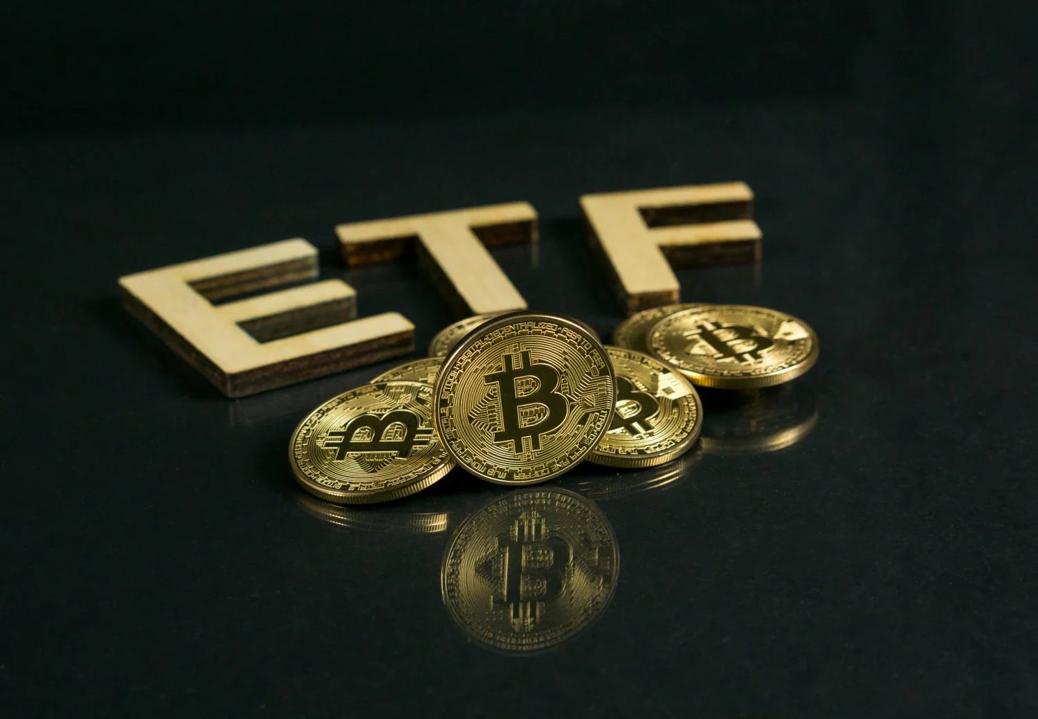 Golden Bitcoin coins are scattered in front of three wooden block letters spelling 'ETF' on a dark, reflective surface. The coins reflect on the surface below, creating a sense of depth and focus on the central Bitcoin coin displaying its 'B' logo.