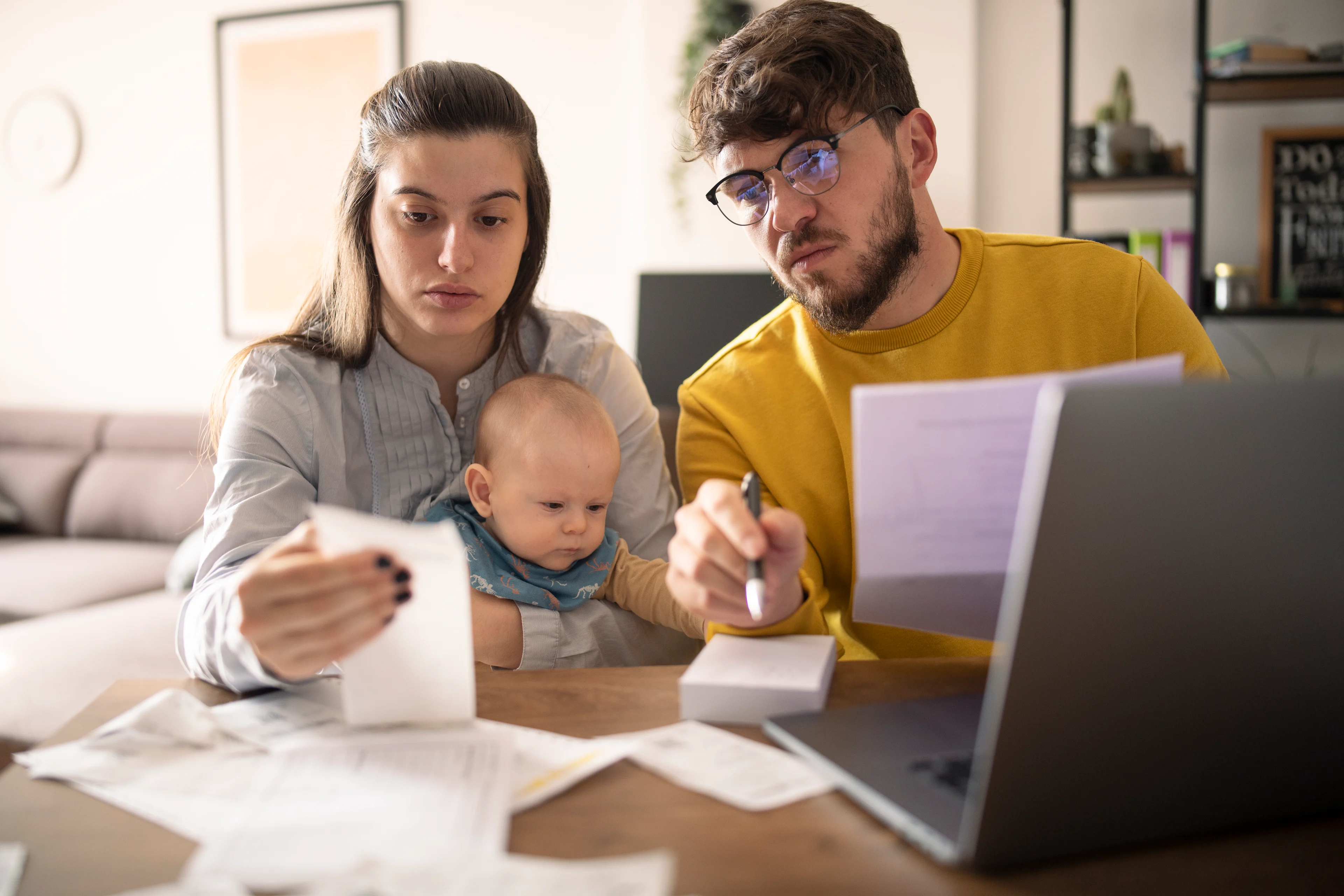 A couple at a kitchen table, with a baby, looking at their finances with concerned looks on their faces. 